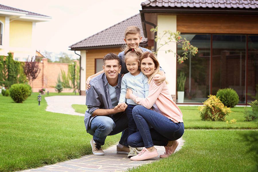Personal Insurance - Young Mother, Father, Son and Daughter Crouch Together Smiling Outside of a Large Home With a Manicured Lawn and Paved Walkway