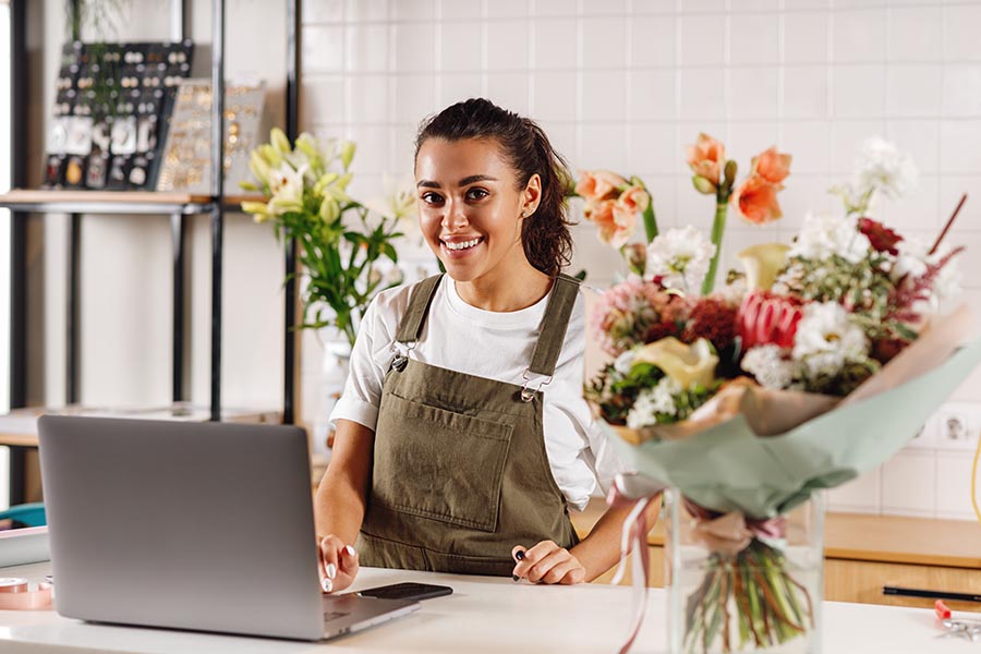 Business Insurance - Flower Shop Owner in Green Overalls Smiles Behind White Counter Using a Laptop, Surrounded by Floral Arrangements and Tools